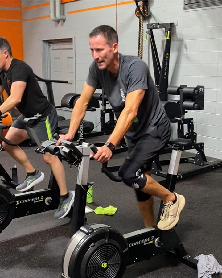 Two men are working out on stationary exercise bikes in a gym. The man in the foreground wears a gray t-shirt, black shorts, and sneakers, and appears focused. Gym equipment is visible in the background.