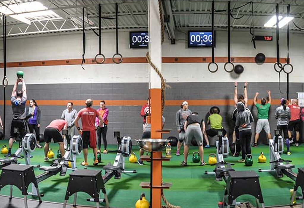 A group of people exercise in a gym, lifting kettlebells and tossing medicine balls against a wall. Rowing machines are lined up in the foreground. Digital timers and gymnastic rings hang on the wall.