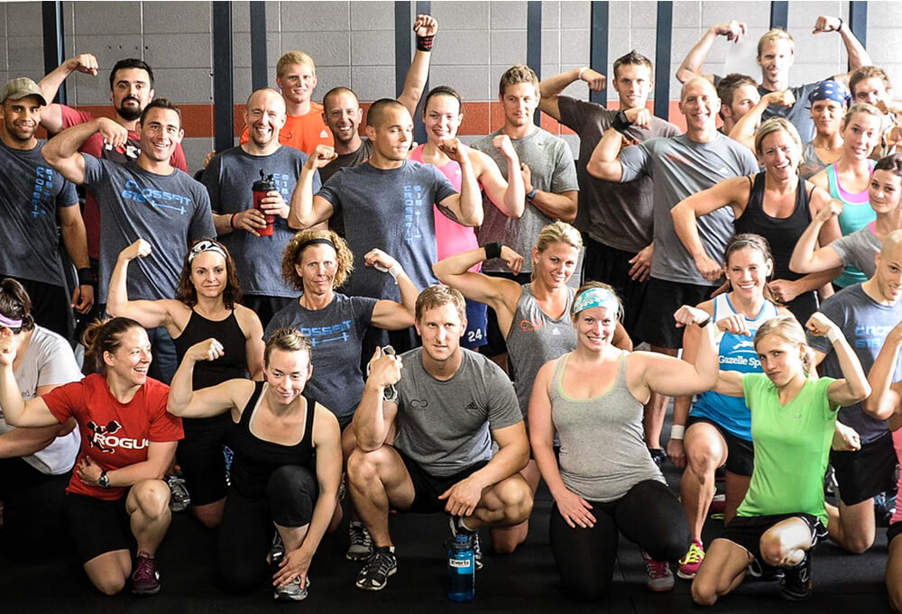 A large group of people in athletic attire pose indoors, smiling and flexing their arms to show their muscles, suggesting a fitness or CrossFit class.