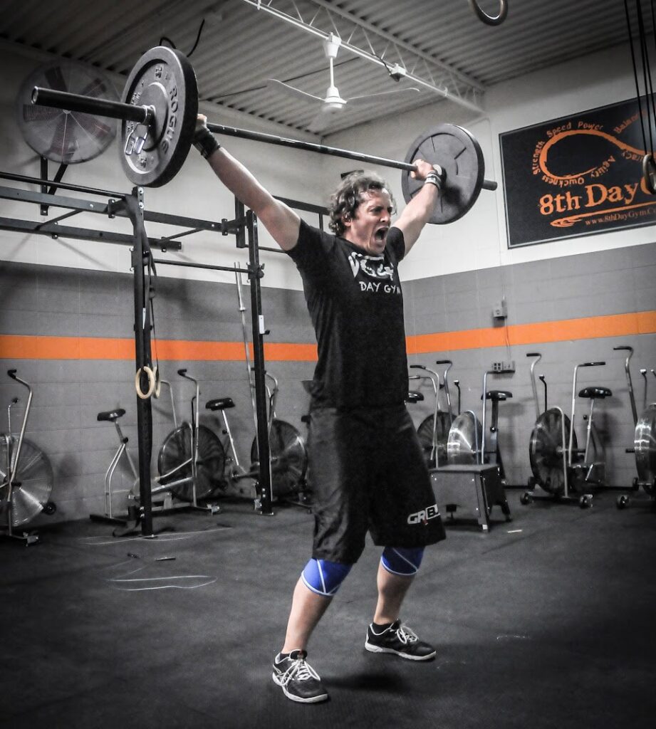 A person in a gym lifts a barbell overhead with both arms extended, appearing to shout in effort. They wear a black shirt, shorts, blue knee sleeves, and athletic shoes. Exercise bikes line the wall in the background.