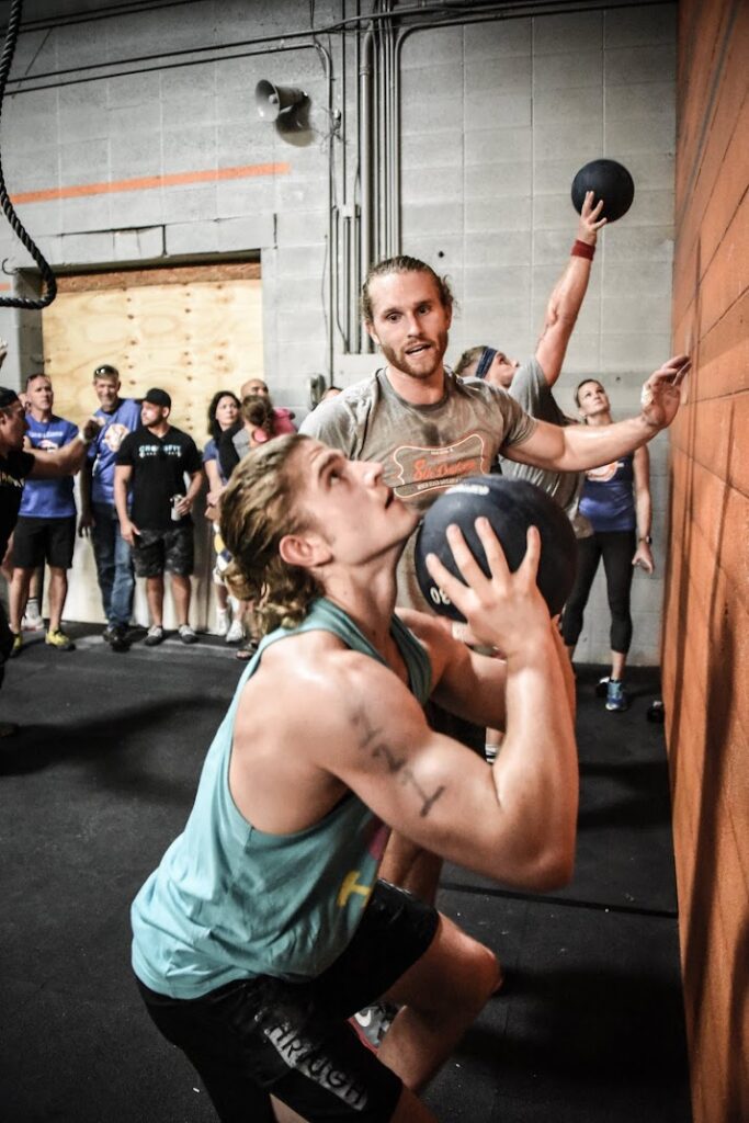 Two men perform wall ball exercises in a gym, each throwing a medicine ball against an orange wall. People stand in the background watching the event. The scene appears energetic and focused.
