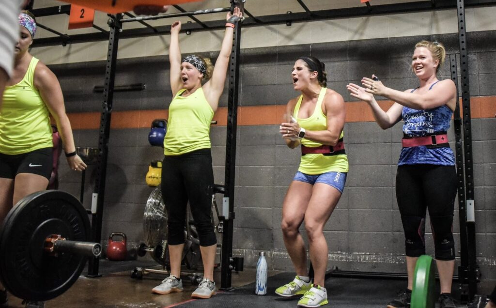 Four women at a gym celebrate a weightlifting achievement. Two are cheering, one raises her arms in victory, and another claps, all in athletic attire next to barbells and kettlebells against a gray wall.