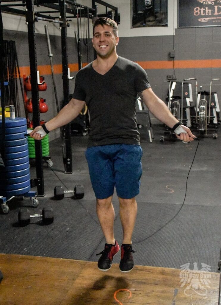 A man in a gym smiles while jumping rope. He wears a dark t-shirt, blue shorts, and black sneakers. Gym equipment and weights are visible in the background.