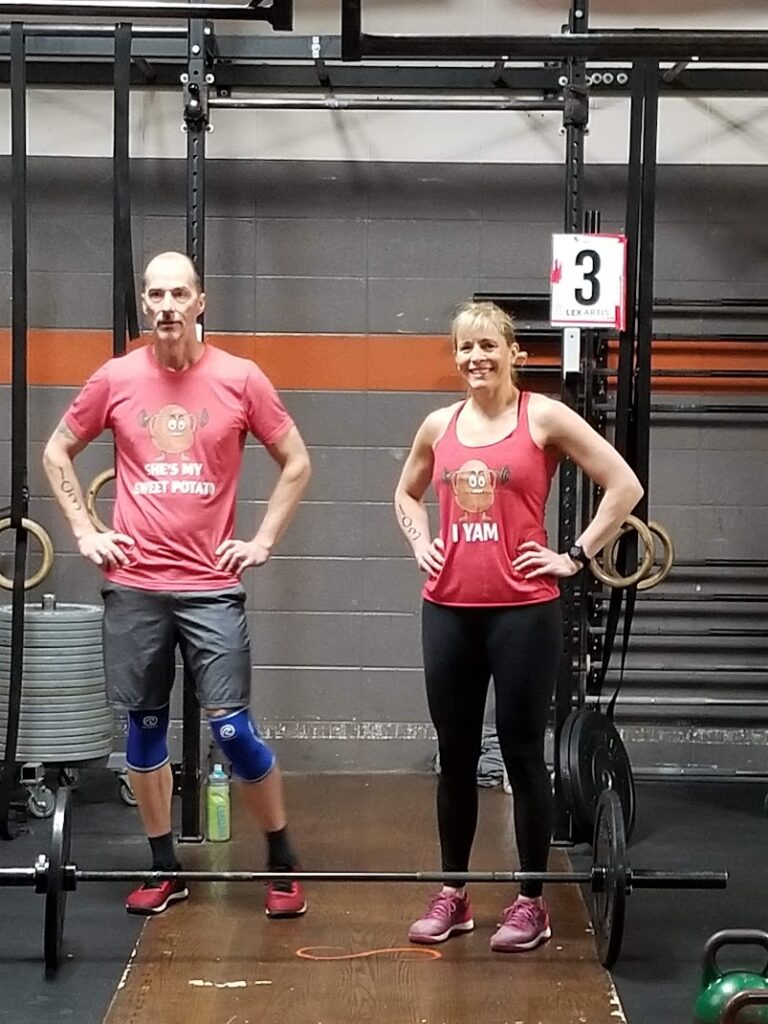 A man and a woman stand side by side in a gym, both wearing matching red tank tops with a cartoon potato and puns. They are in front of gym rings and a barbell, smiling and ready to work out.