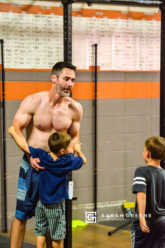 A shirtless man stands in a gym, smiling as two young boys interact with him. Workout routines are listed on a whiteboard in the background, and gym equipment is visible around them.