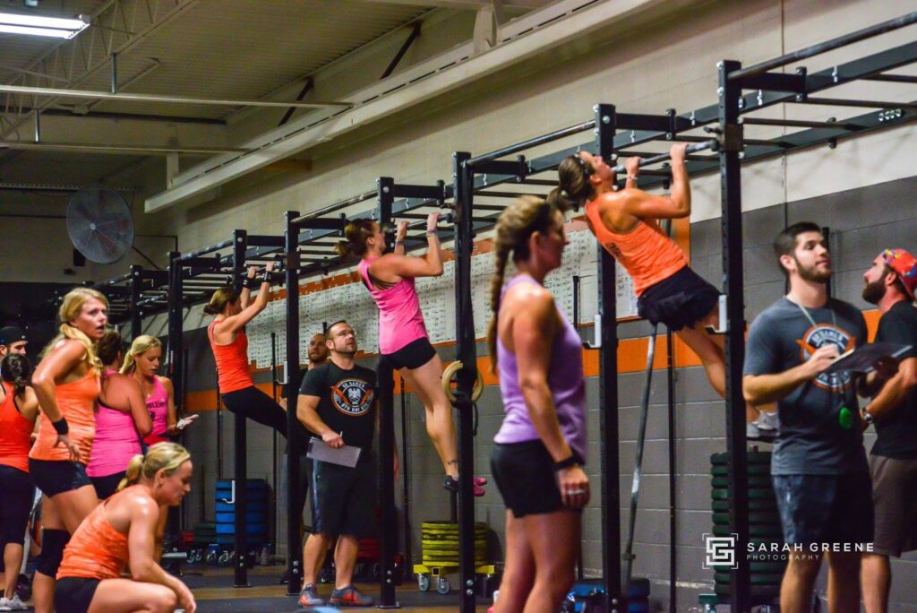 A group of women in athletic wear perform pull-ups in a gym while others watch or rest nearby. Several people, including judges with clipboards, observe the activity. The gym has high ceilings and workout equipment.