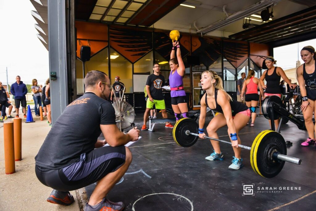 A coach kneels and observes as a woman lifts a barbell in a gym. Other people are working out in the background, including a woman lifting a kettlebell overhead. The scene is energetic and focused.