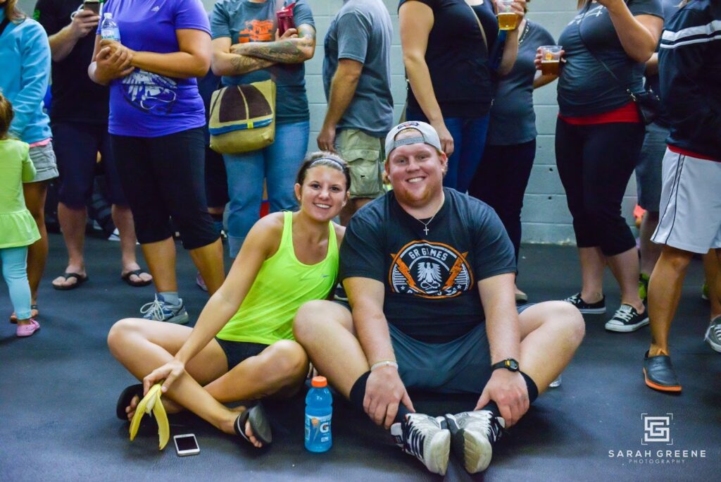 A smiling man and woman sit on the gym floor, surrounded by people standing. The woman holds a banana, and a sports drink sits in front of them. Both are casually dressed in athletic wear.