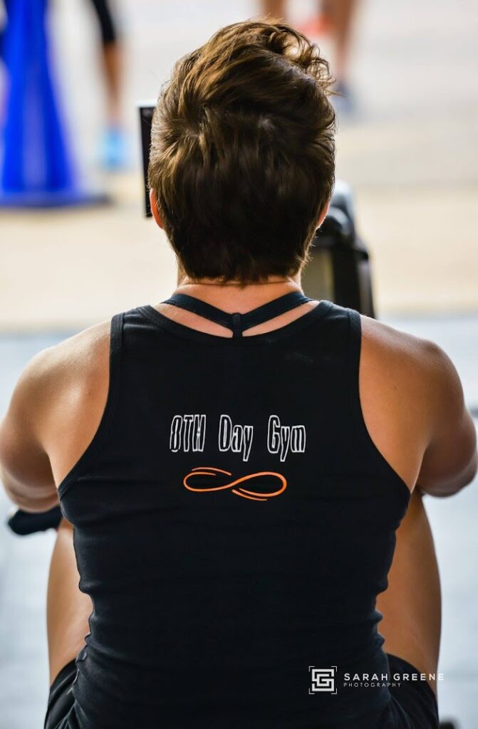A person with short hair, viewed from behind, wears a black tank top with 8TH Day Gym printed on the back while using gym equipment. The photo is by Sarah Greene Photography.