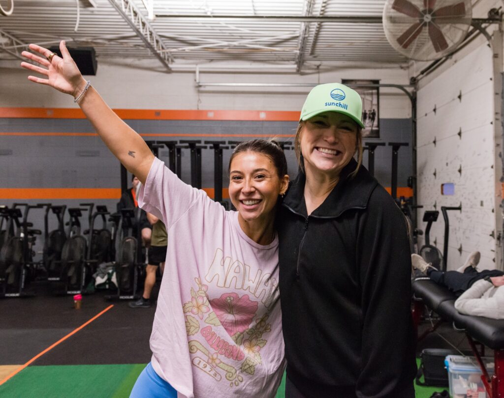 Two women smiling in a gym. One wears a pink graphic t-shirt with an arm raised; the other wears a black jacket and green cap. Exercise equipment and people are visible in the background.