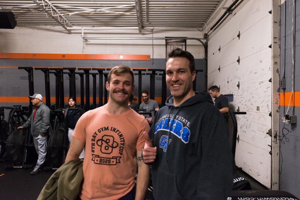 Two men smiling at a gym; one wears a peach T-shirt with a gym logo, the other wears a dark sweatshirt with blue text. Several people exercise in the background next to gym equipment and a white wall.