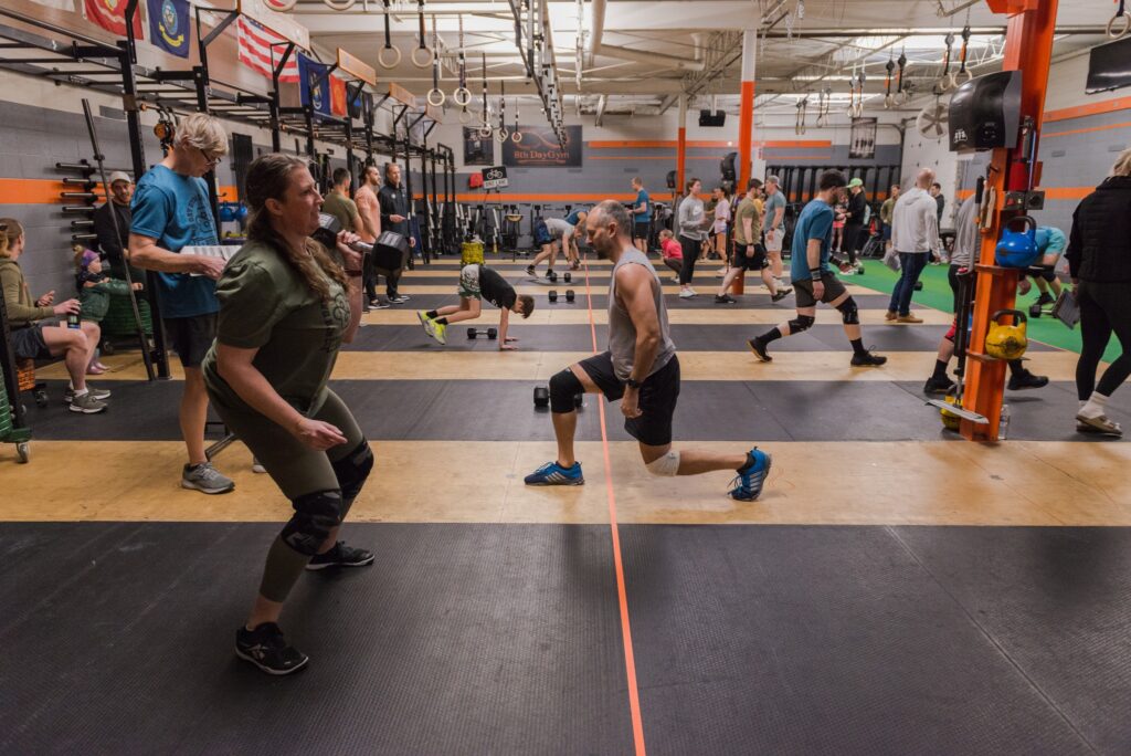 A group of people exercise in a gym, performing various strength and cardio workouts. Two people in the foreground do lunges with dumbbells, while others use equipment or train in the background.