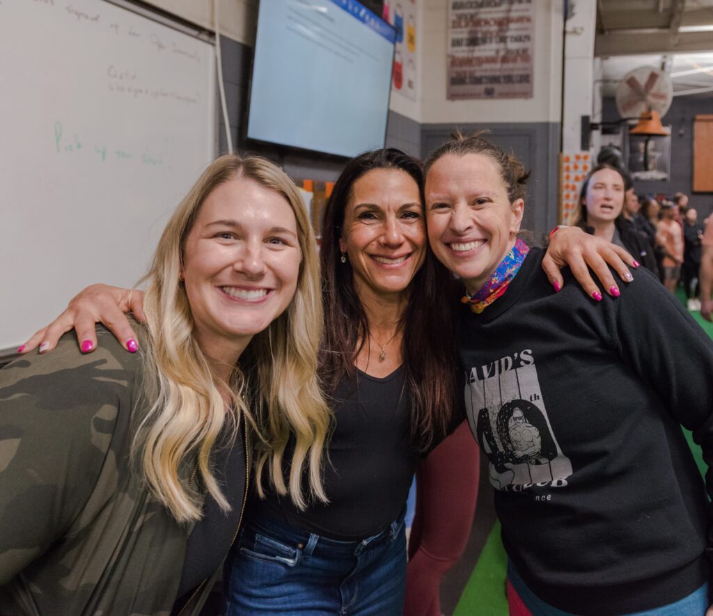Three women are smiling and posing together indoors at what appears to be a gym or event, with people and a whiteboard in the background.