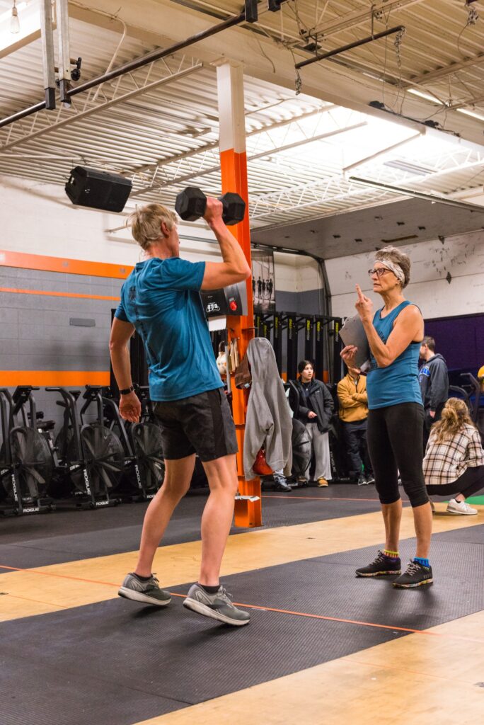 A young person lifts a dumbbell overhead while a woman, possibly a coach, gestures and gives instructions in a gym with other people and equipment in the background.