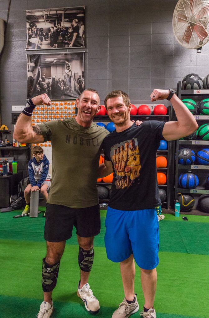 Two men pose smiling and flexing their arms in a gym, with colorful medicine balls on shelves behind them. One wears a green shirt and black shorts, the other a black graphic t-shirt and blue shorts. Other gym-goers sit in the background.