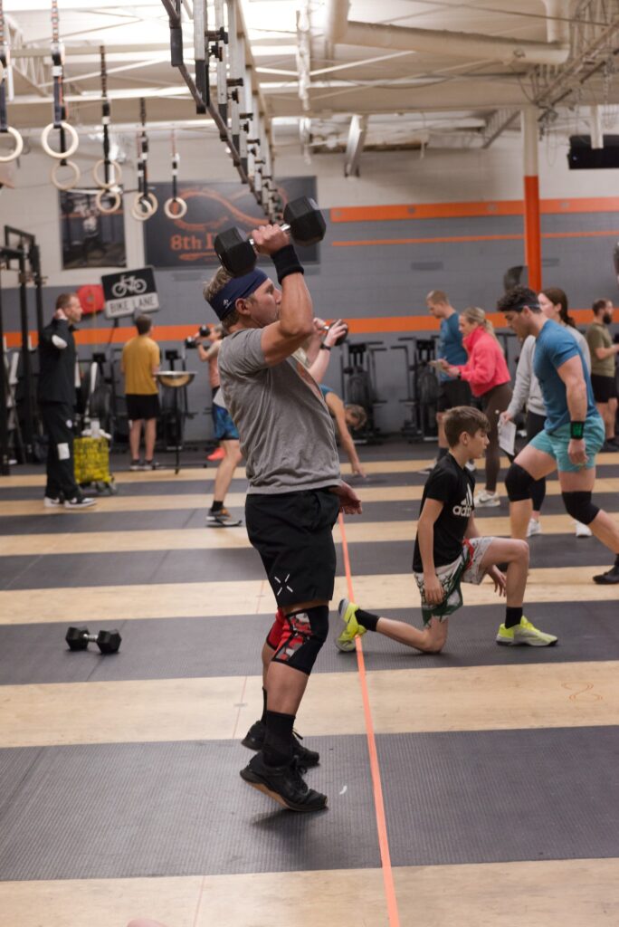 A man lifts a dumbbell overhead in a gym, surrounded by others exercising. Some people are lunging, while others prepare equipment. The gym has exposed ceilings, exercise rings, and orange and gray walls.