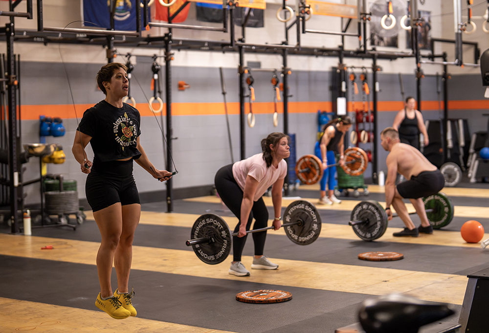 People working out in a gym: one person is jump roping in the foreground, while others behind them are lifting barbells. Various gym equipment and weights are visible in the background.
