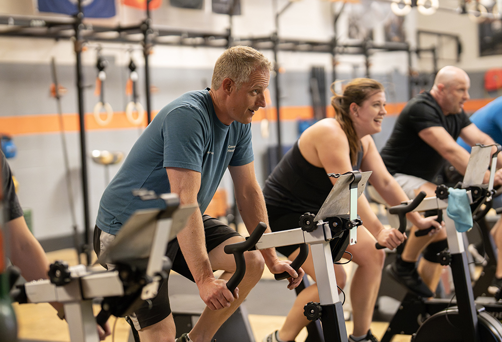 A group of adults exercise on stationary bikes in a gym, focusing on their workout. The gym equipment and fitness gear are visible in the background.