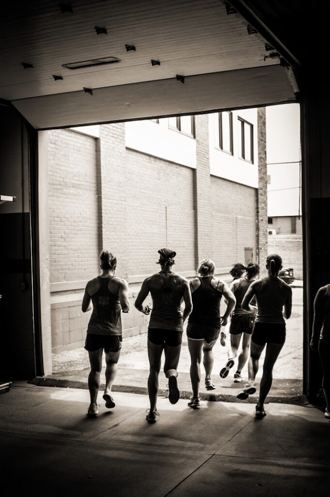 Five people in athletic wear run out of a dimly lit gym through an open garage door into a sunlit alleyway, captured in black and white.