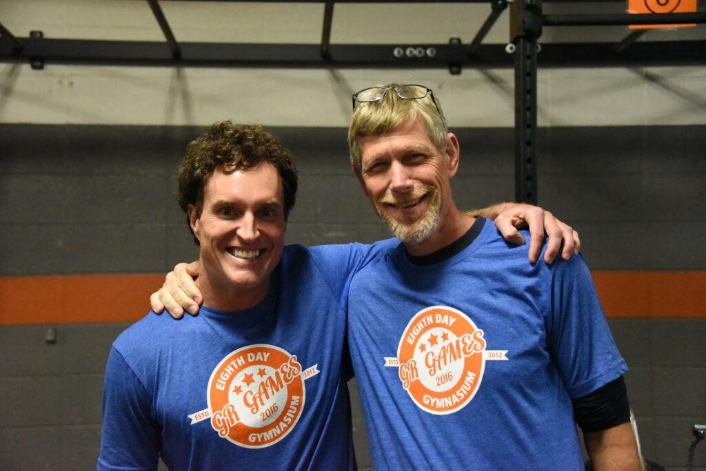 Two men smiling and standing side by side with their arms around each other, both wearing matching blue Eighth Day Games Gymnastics t-shirts in a gym setting.