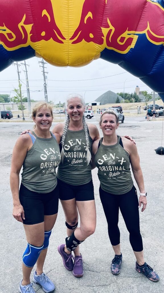 Three women in matching workout outfits and knee braces stand smiling under a large inflatable Red Bull archway outdoors, posing after a fitness event.