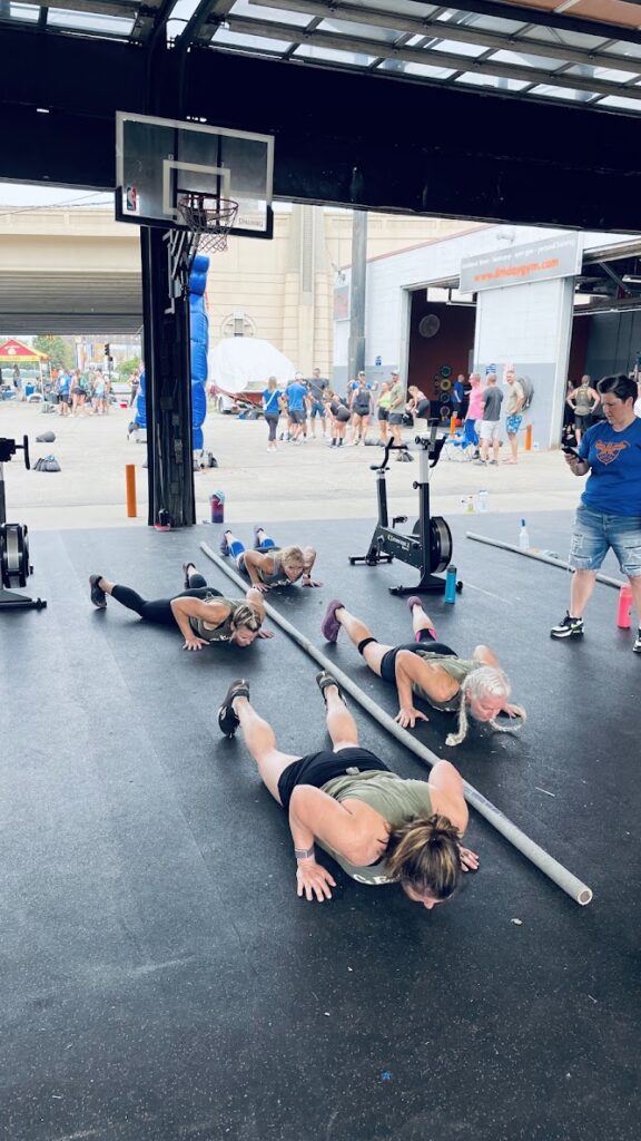 Four women perform push-ups in a gym, facing a metal bar on the floor, while others watch or participate. The gyms large open door reveals a crowd and outdoor event in the background.