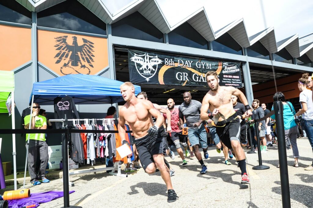 A group of competitors, some shirtless, sprint from the starting line outside a gym during a fitness event. Spectators and tents are nearby, with a banner reading “8th Day Gym GR Games” in the background.