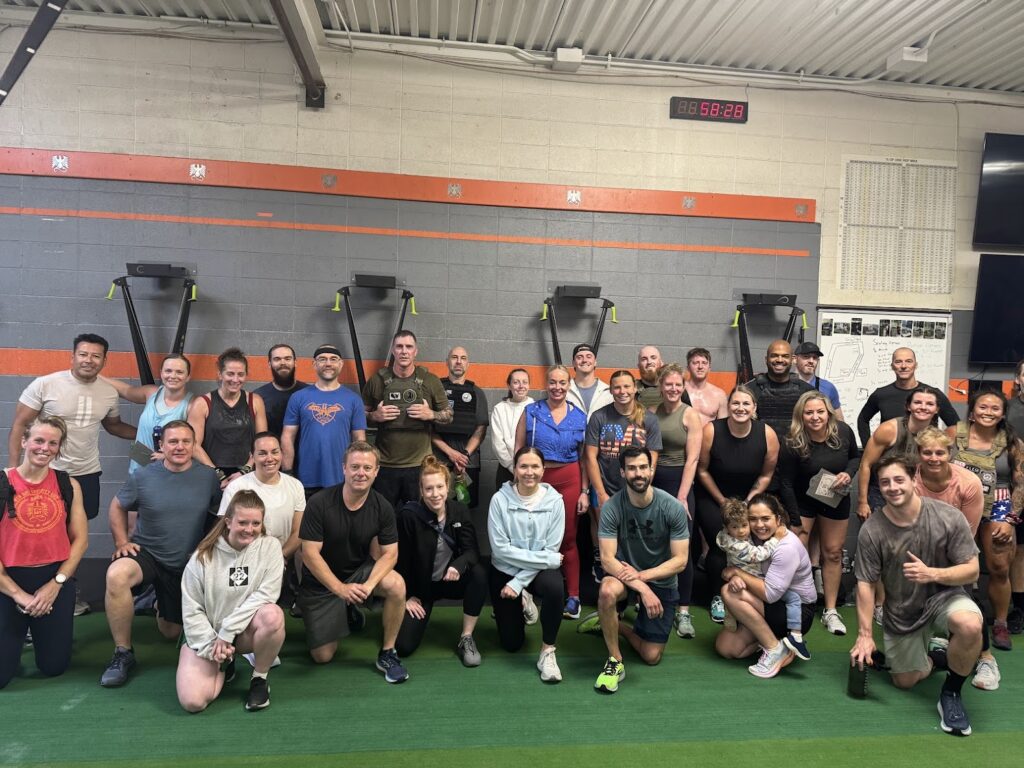 A large group of people pose and smile together inside a gym, standing and kneeling on green turf with workout equipment and wall timers visible in the background.