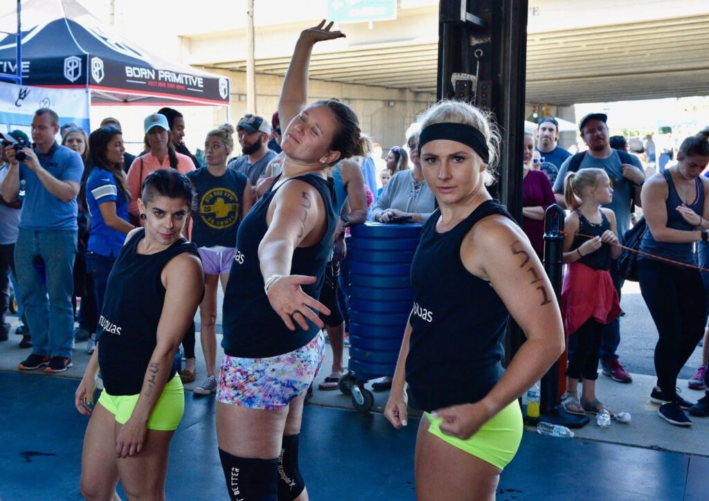 Three women in matching black tank tops and neon yellow shorts pose confidently at a fitness event, with a crowd of spectators standing behind them and various tents and banners visible.