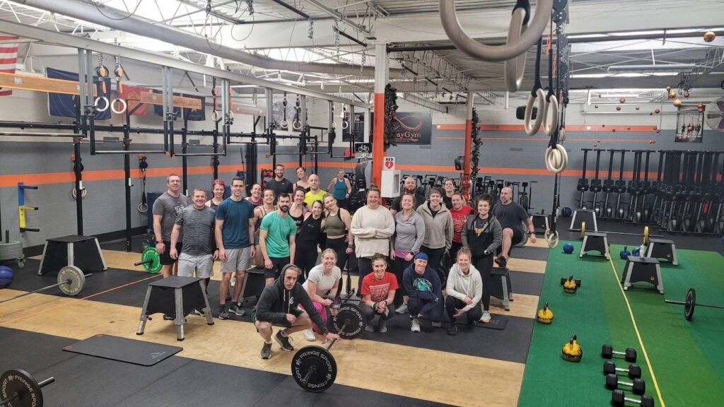 A group of people pose and smile for a photo in a large gym with workout equipment, weights, and turf. Gymnastic rings hang from the ceiling, and kettlebells and barbells are on the floor nearby.