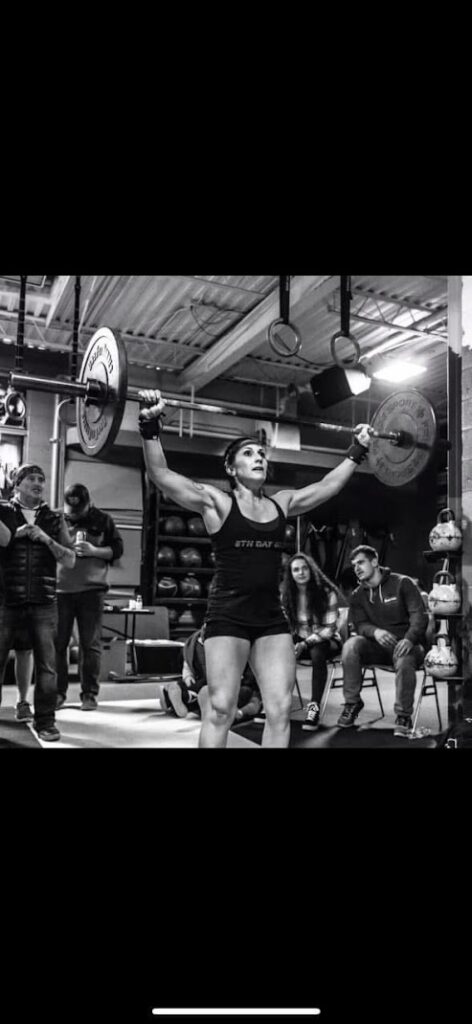 A woman lifts a barbell overhead in a gym, surrounded by people watching her. The image is in black and white, capturing the intense focus and strength of the lifter in a competition or training setting.