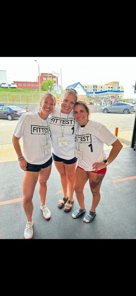 Three smiling women stand close together indoors, each wearing white FITTEST T-shirts and event name tags. They appear happy, posing for the photo, with buildings and a street visible through the windows behind them.