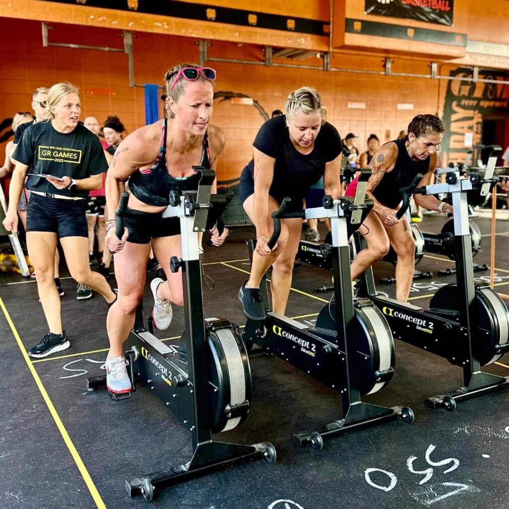Four women intensely compete on stationary bikes at an indoor fitness event. They lean forward, focused and determined, with other participants and spectators visible in the background.