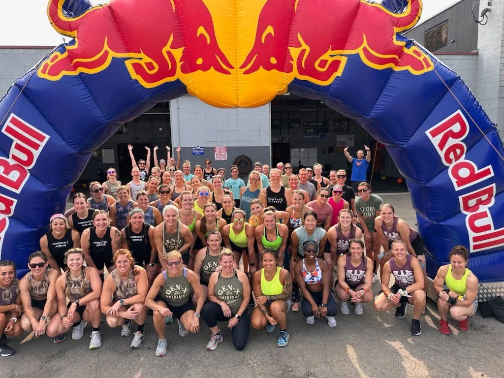 A large group of people in athletic wear pose and smile together under an inflatable Red Bull arch at an outdoor event. The group appears to be diverse and energetic.