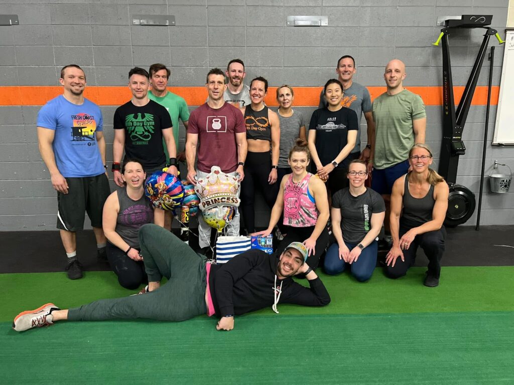 A group of 15 people pose and smile in a gym, with fitness equipment in the background. One person is lying on the floor in front, and others are holding balloons and a gift bag.