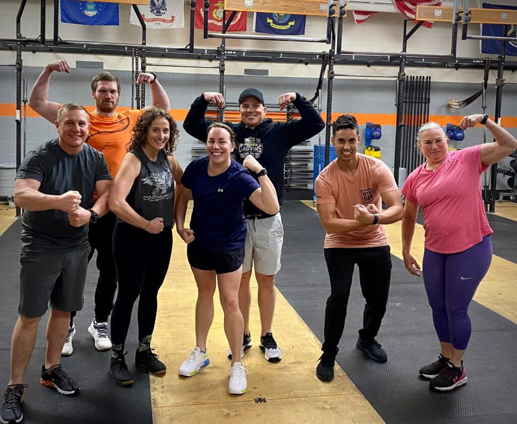 A group of seven adults at a gym pose together, smiling and flexing their arm muscles. They stand on black mats with gym equipment and flags visible in the background.
