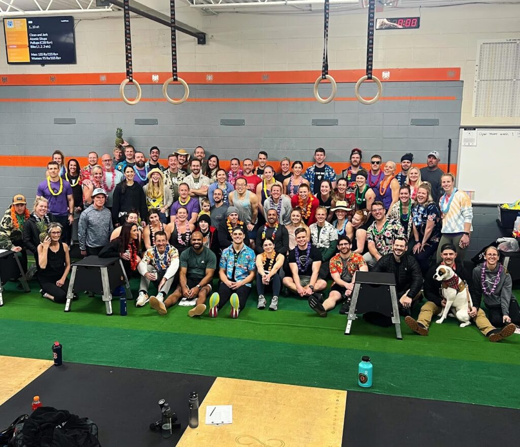 A large group of people in colorful, tropical-themed clothing and leis pose and smile together in a gym, surrounded by workout equipment and gymnastics rings.