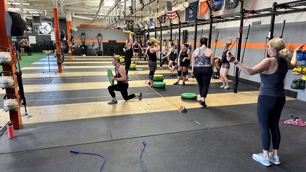 A group of people participate in a CrossFit class in a gym. Some do lunges with weights, while others jump rope. The gym has equipment, weights, and flags on the wall. The atmosphere is active and energetic.