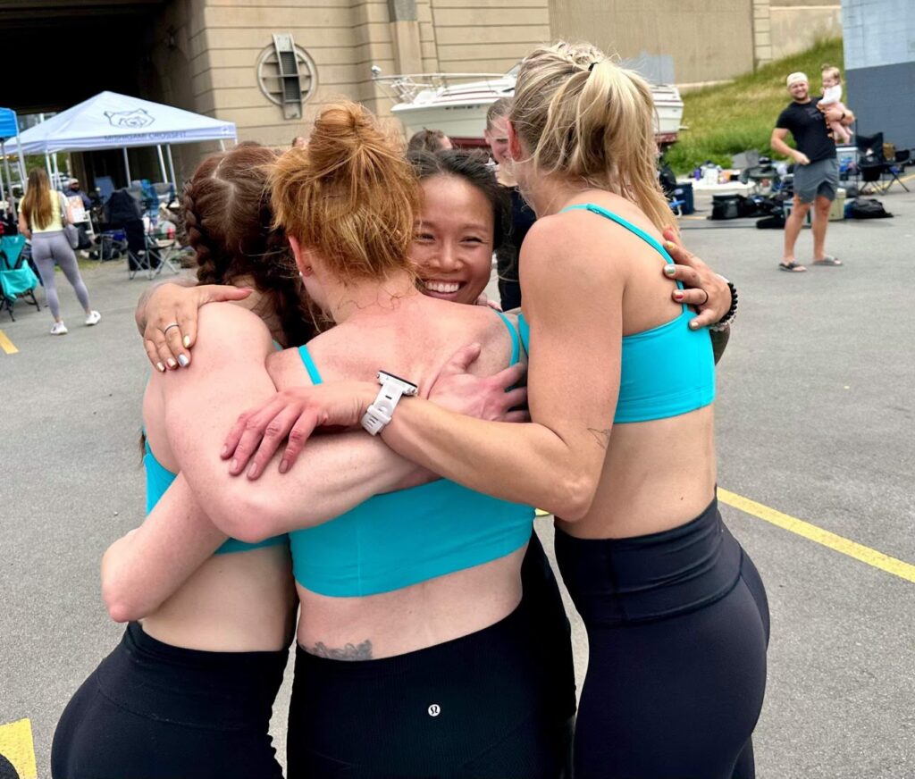 Four women in athletic wear, smiling and embracing in a group hug outdoors, with tents and people visible in the background. They appear happy and supportive, possibly after a fitness event or competition.