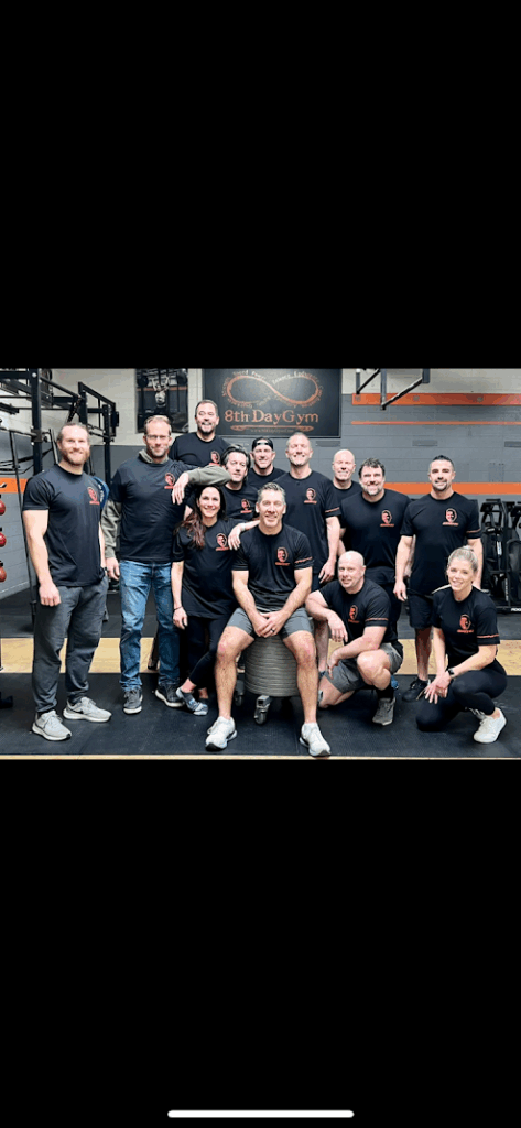 A group of people in matching black shirts pose together and smile inside a gym, with workout equipment visible in the background and a gym logo on the wall behind them.