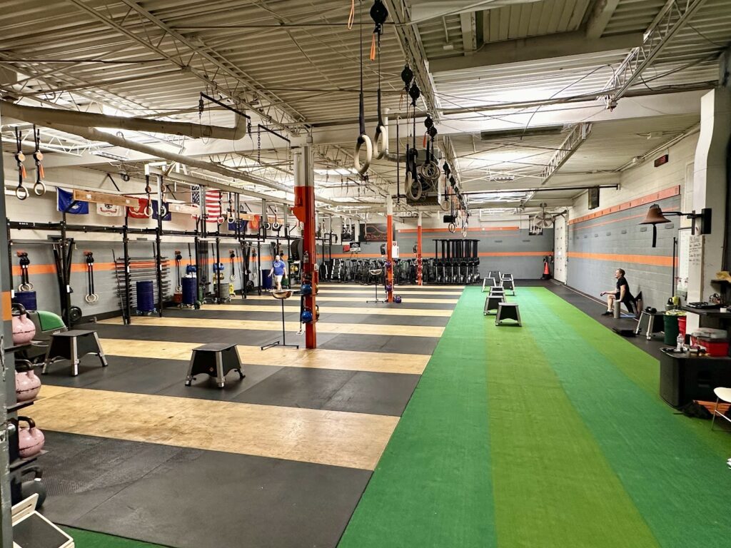 A spacious gym with rubber flooring, green turf track, various exercise equipment, kettlebells, pull-up bars, gymnastic rings, and benches. An American flag and other banners hang on the wall; a person sits on a bench near the side.
