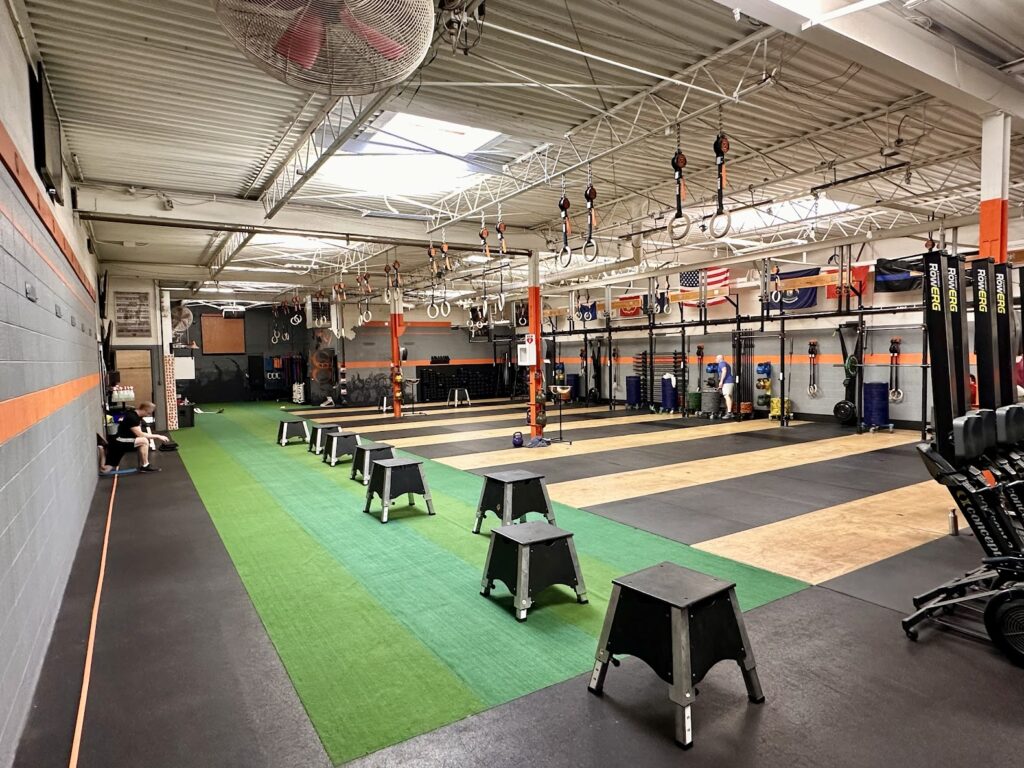 A spacious gym with turf and rubber flooring, plyometric boxes lined up, pull-up bars, gymnastic rings, rowing machines, and various fitness equipment. Flags hang on the back wall; a few people are working out.