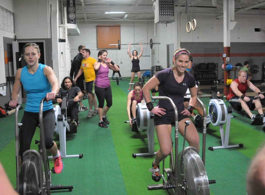 A group of people exercise in a gym; some pedal on stationary bikes, others use rowing machines, and a woman in the back lifts a weighted bar overhead. The atmosphere is energetic and active.