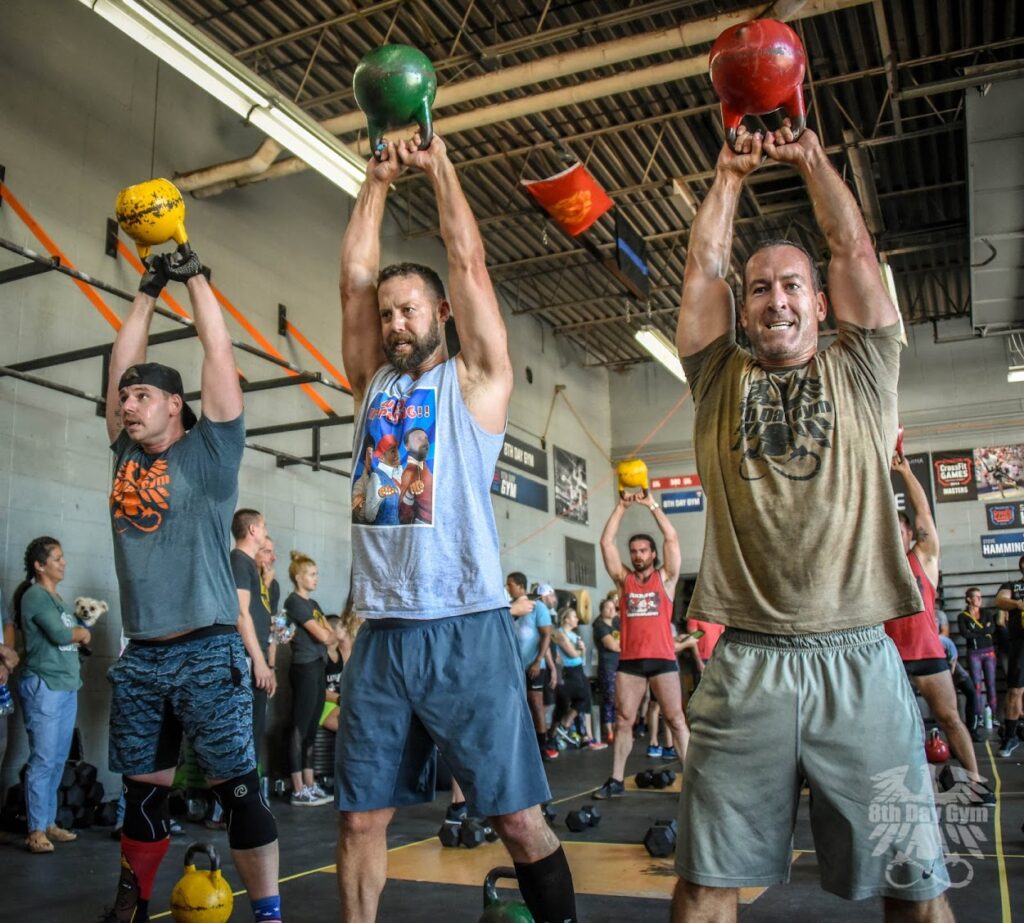 Three men in athletic gear lift kettlebells overhead in a gym as others watch. The scene is energetic, with sunlight streaming in and banners on the walls. The men appear focused and determined during the workout.