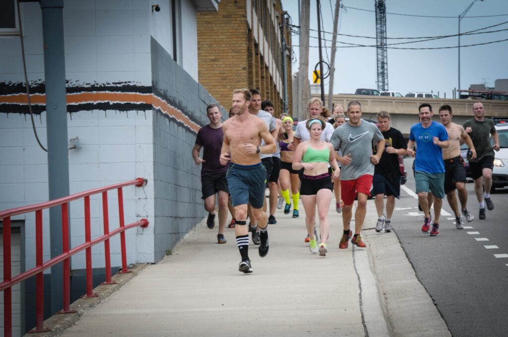 A group of people, including men and women, are running together on a city sidewalk next to a road. Some runners wear athletic gear, while one man in front is shirtless. The background shows buildings and utility poles.