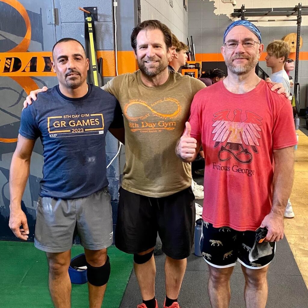 Three men stand together in a gym, smiling at the camera. They are wearing workout clothes, and gym equipment is visible in the background. One man gives a thumbs up, and others are exercising in the background.