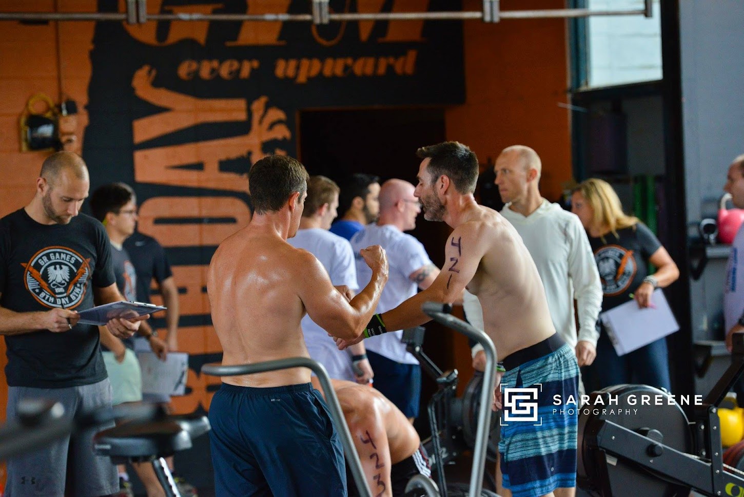 A group of people inside a gym, with two shirtless men in the foreground giving each other a fist bump. Others in the background are holding clipboards and watching. Gym equipment and a large workout-themed mural are visible.